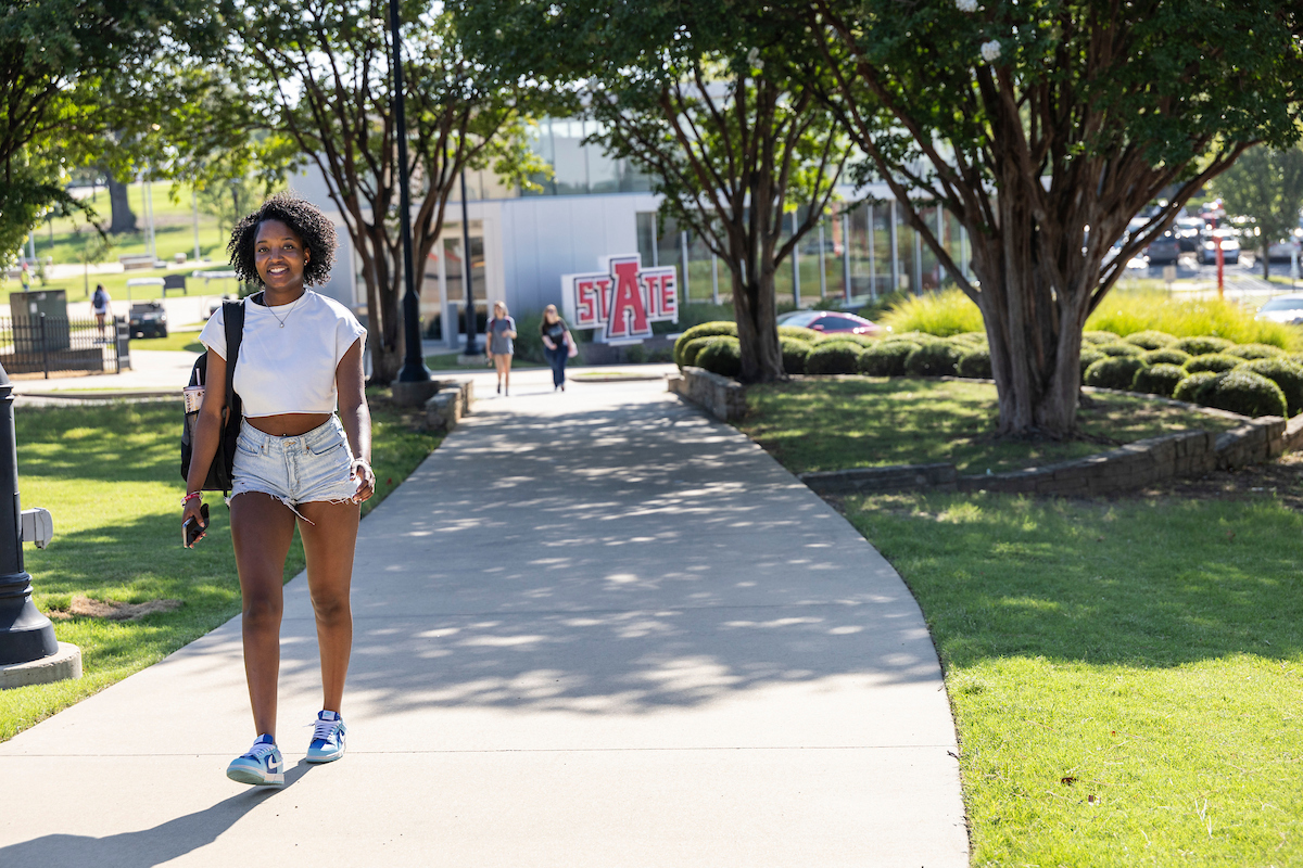 AState student smiles while walking on a shaded campus sidewalk with the A-State Welcome Center in the background