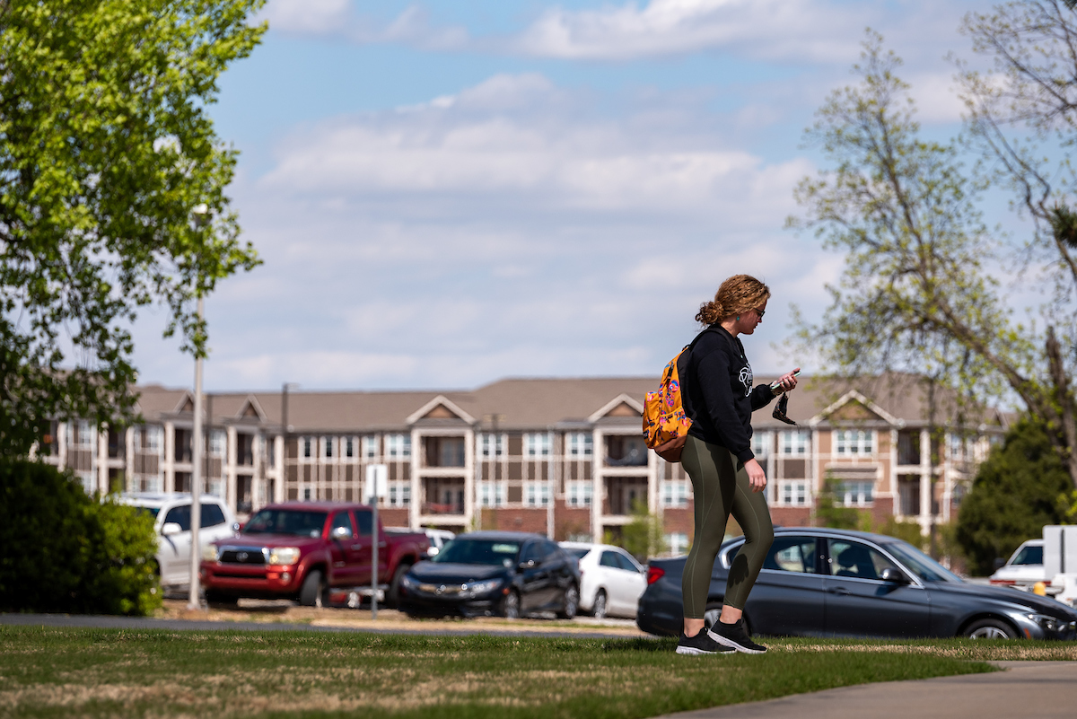 Student walks on campus sidewalk near parked cars with apartments in the background.