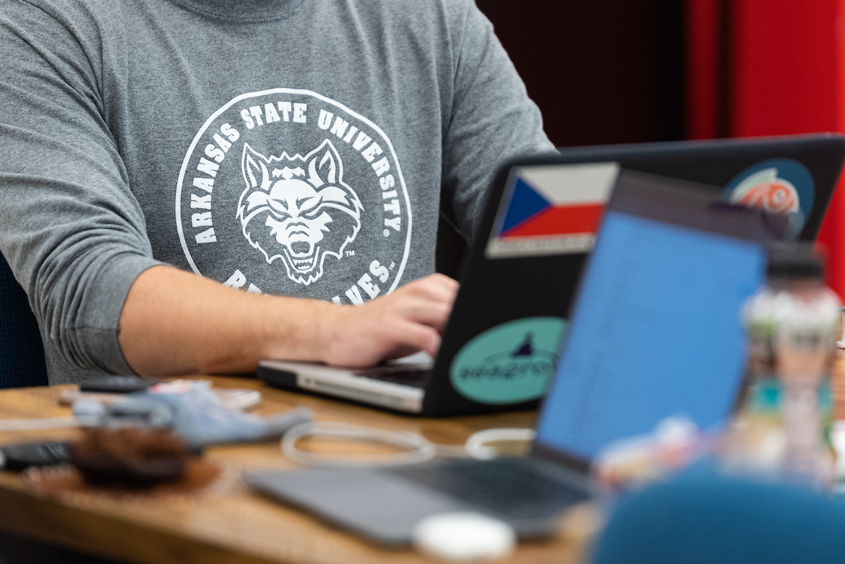 Close-up of A-State student wearing a Red Wolves shirt while working on a laptop at a desk.