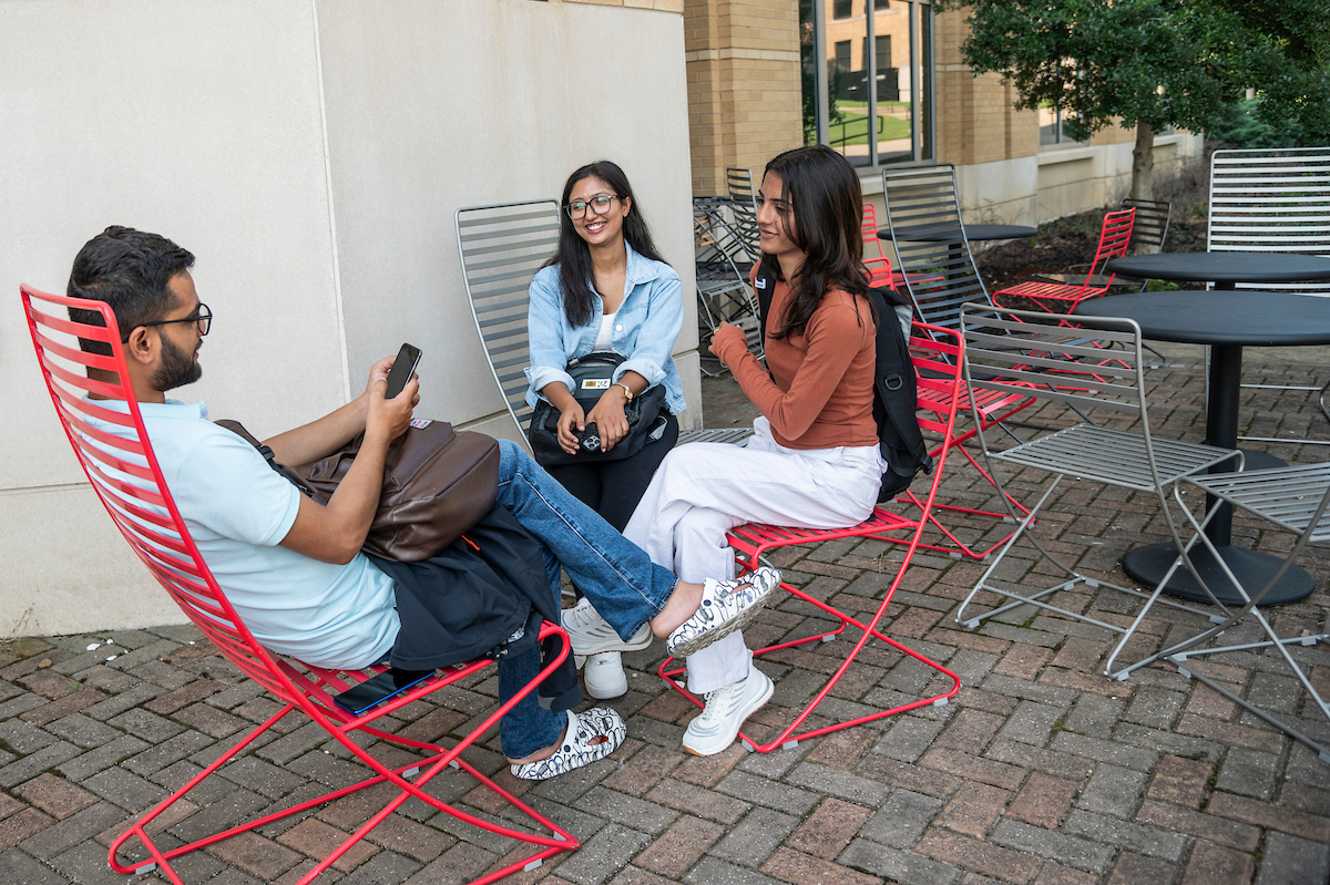 Three students relax and chat on red chairs at an outdoor campus patio, enjoying downtime between classes.