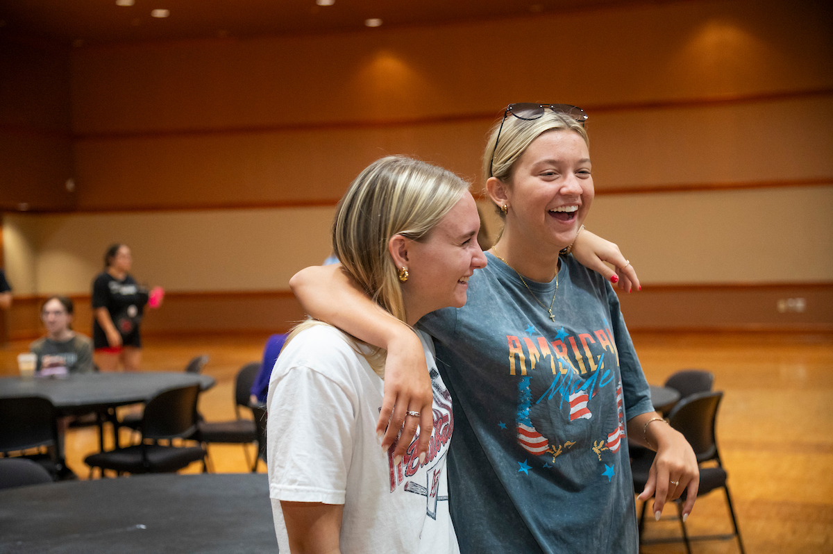 Two smiling astate students with arms around each other laugh during a casual campus event indoors.