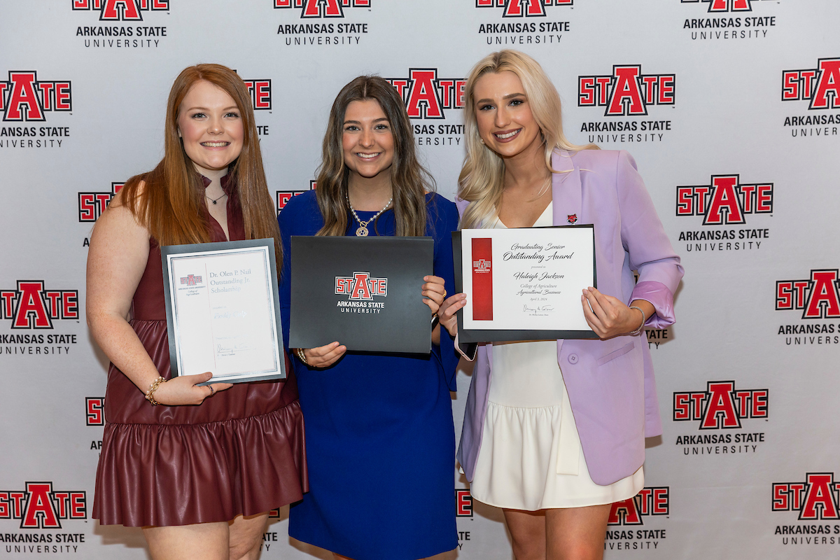 Three A-State students showcase their awards in front of a university backdrop.