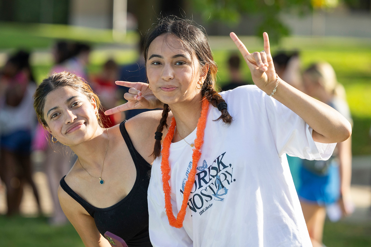 Two A-State students smile and flash peace signs while enjoying a sunny outdoor campus event.