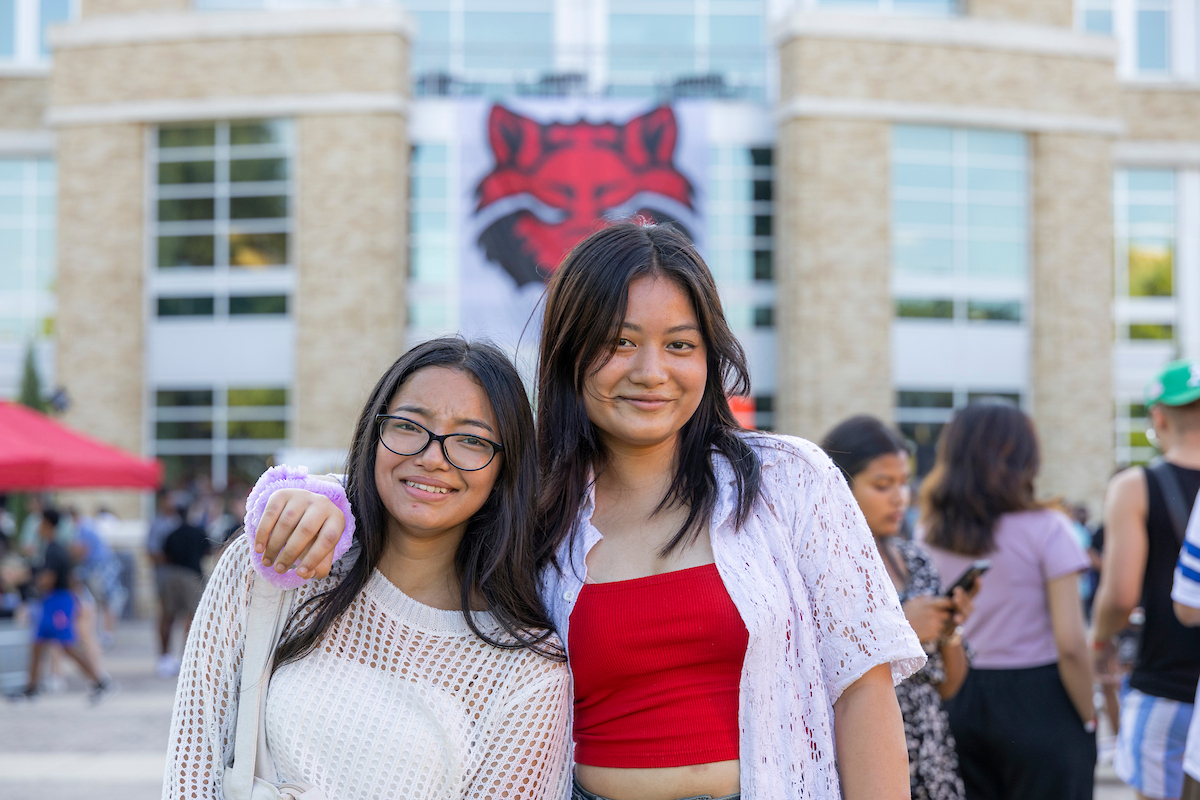 Two A-State students smile in front of a Red Wolves banner during a campus event.