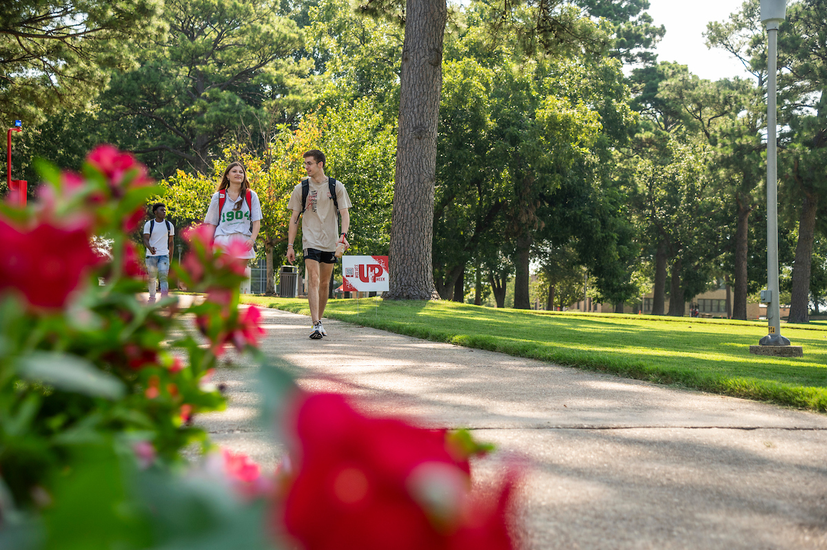 A-State students walk along a tree-lined sidewalk bordered by red flowers on a sunny campus day.