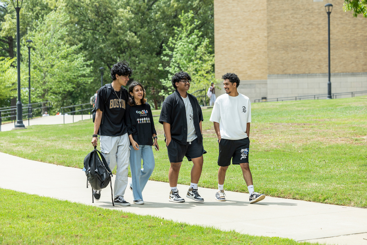Group of A-State students walk and chat on a sunny campus sidewalk surrounded by green trees.