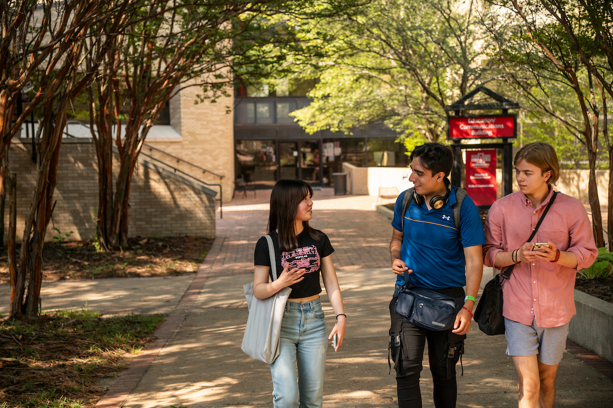 Three students walk and talk in front of the Communication Building on a shaded campus pathway.