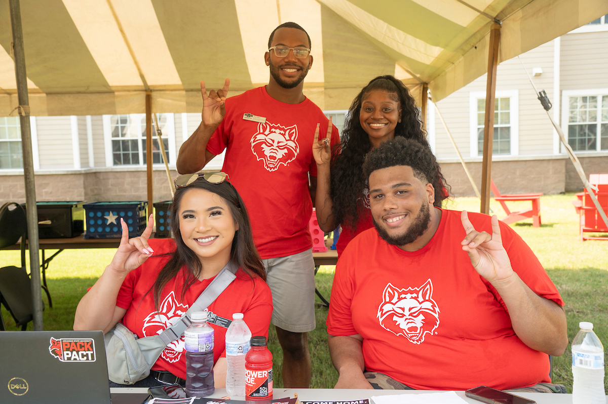 Staff and students wearing Red Wolves shirts smile and flash the “Wolves Up!” sign at a welcome week event under a tent.