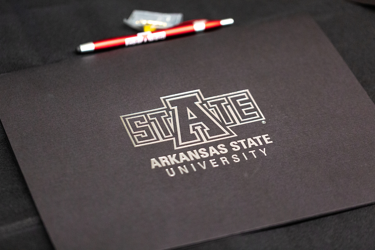 A-State graduation folder with silver writing on a black table and pen.
