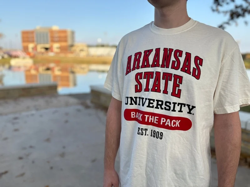 A person wearing an Arkansas State University “Back the Pack” T-shirt stands near a campus pond with Centennial Bank Stadium visible in the background on a clear day.