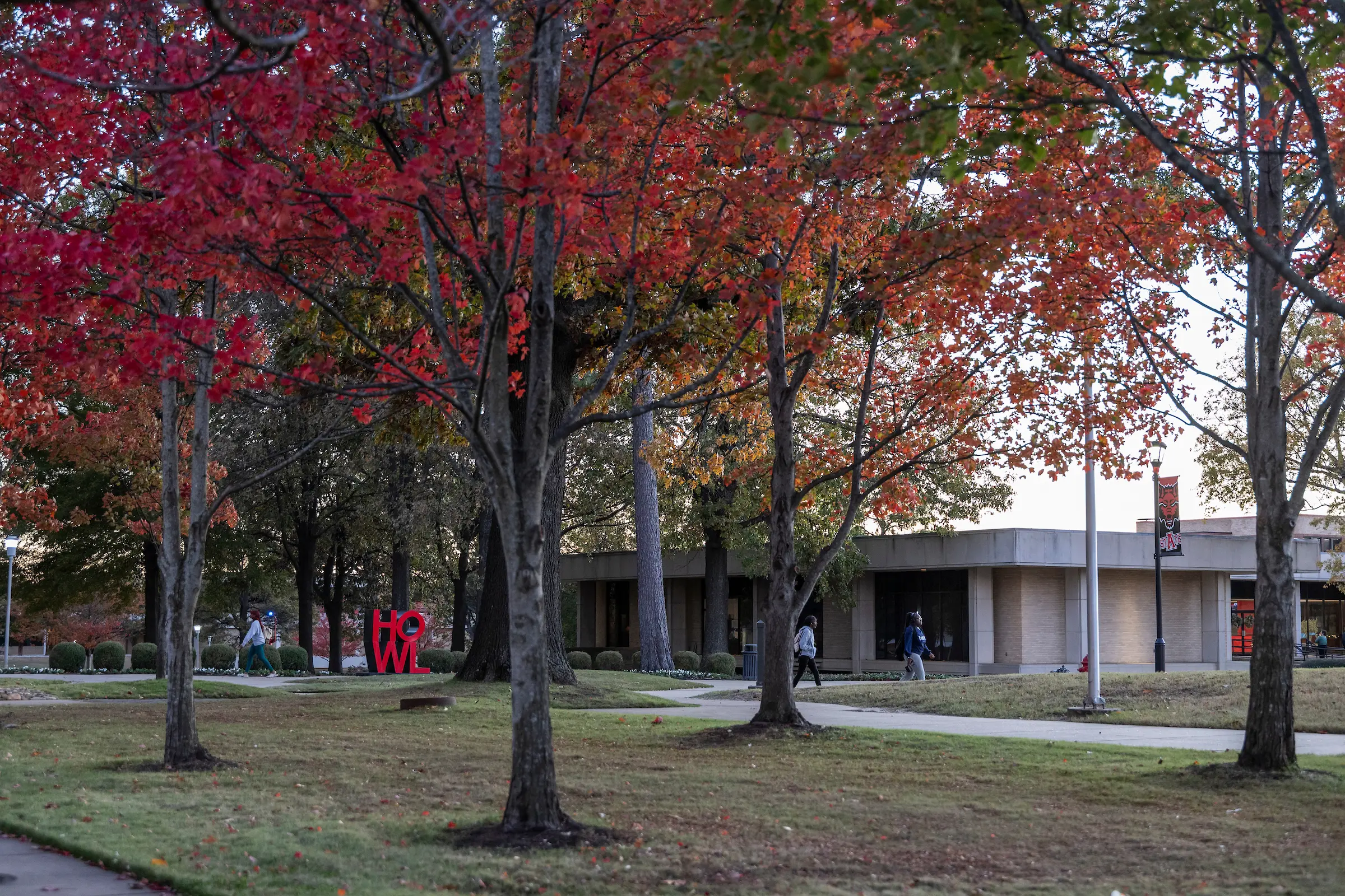 Red and orange fall leaves frame a quiet campus sidewalk near the large red HOWL sculpture