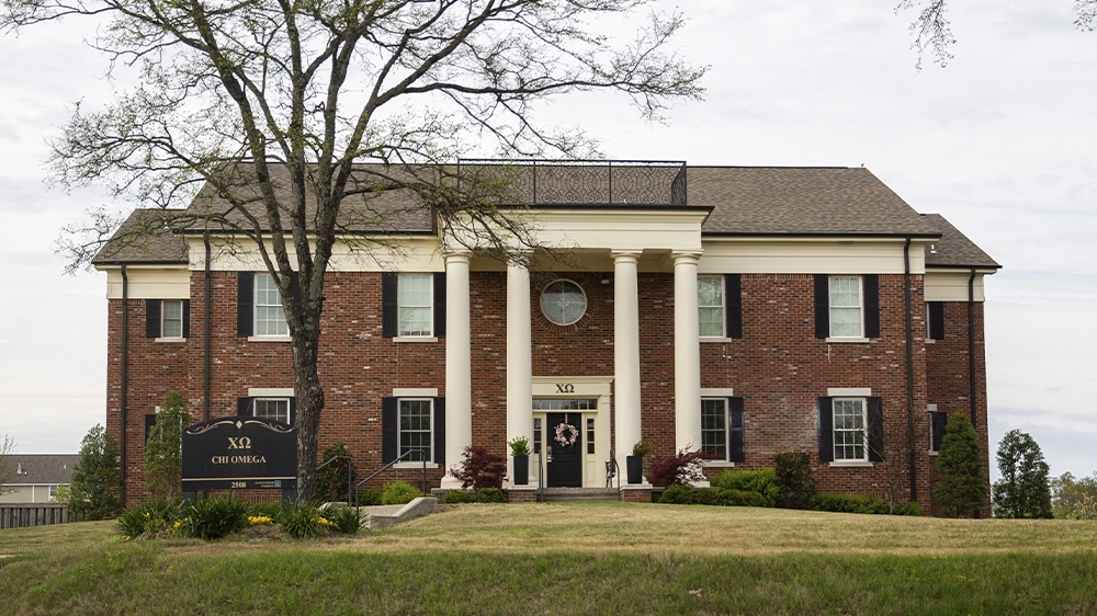 Two-story red brick sorority house with white columns and black shutters. A black sign in front reads “ΧΩ Chi Omega.” The house sits on a grassy lawn with a tree and shrubs near the entrance.