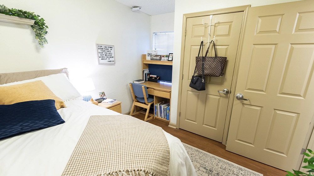 A bedroom with a white bedspread, beige throw, and colorful pillows. A desk with books and decor sits near the window, and a closet door is visible.