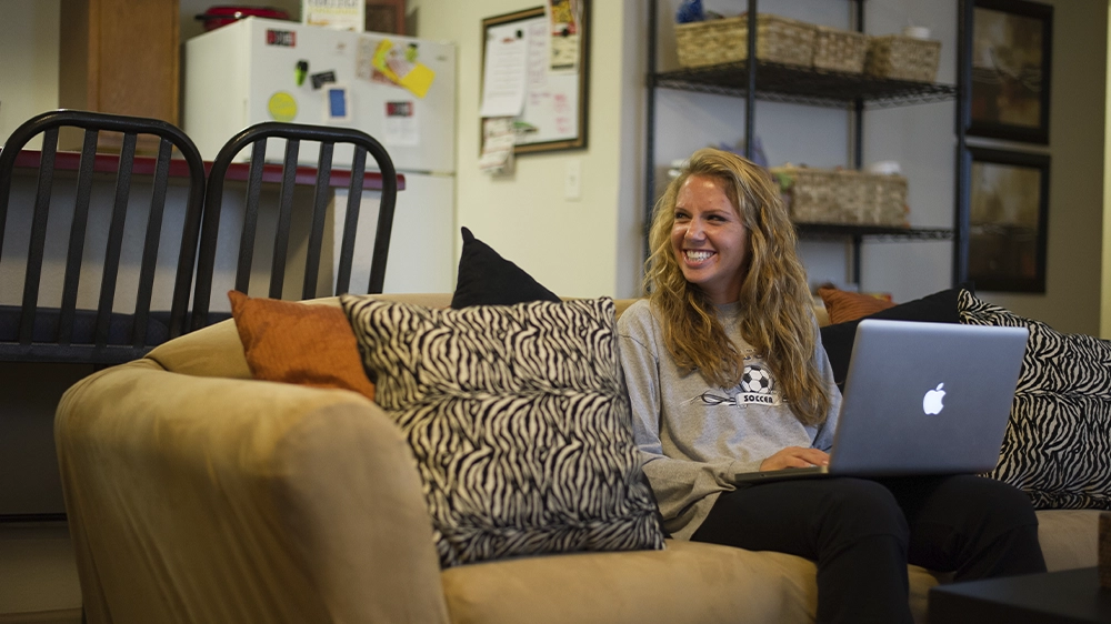A living area with a beige sofa and patterned pillows, adjacent to a kitchen with a breakfast bar and stools. Shelving with baskets adds storage.