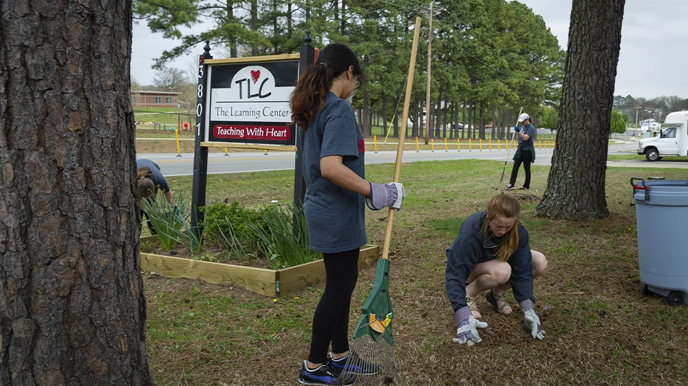 Two individuals working outdoors near a sign that reads “The Learning Center – Teaching With Heart,” raking and cleaning up landscaping.