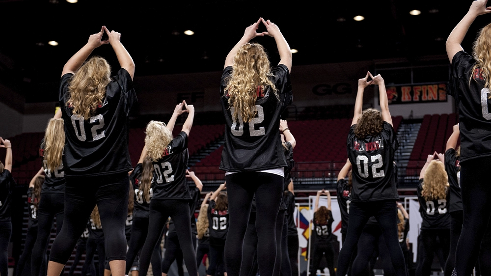 Group of performers in black jerseys with “02” on the back forming hand signs during a choreographed routine on stage.