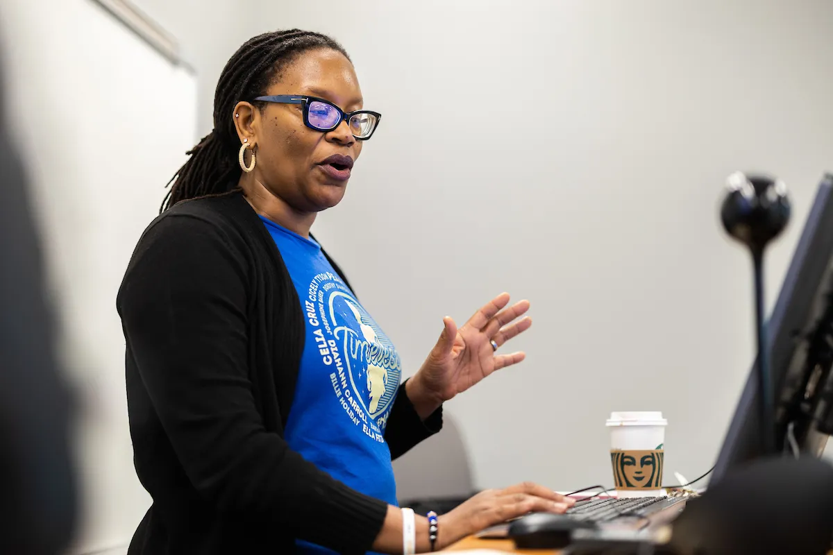 A professor teaches in front of a computer while speaking and using hand gestures.