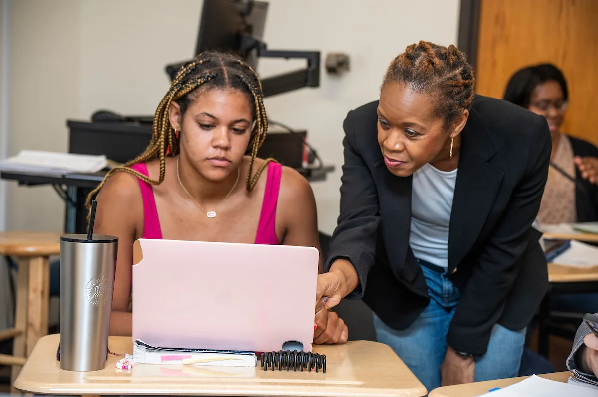 An A-State faculty member mentors a student at desk, pointing at laptop screen in a supportive classroom setting.