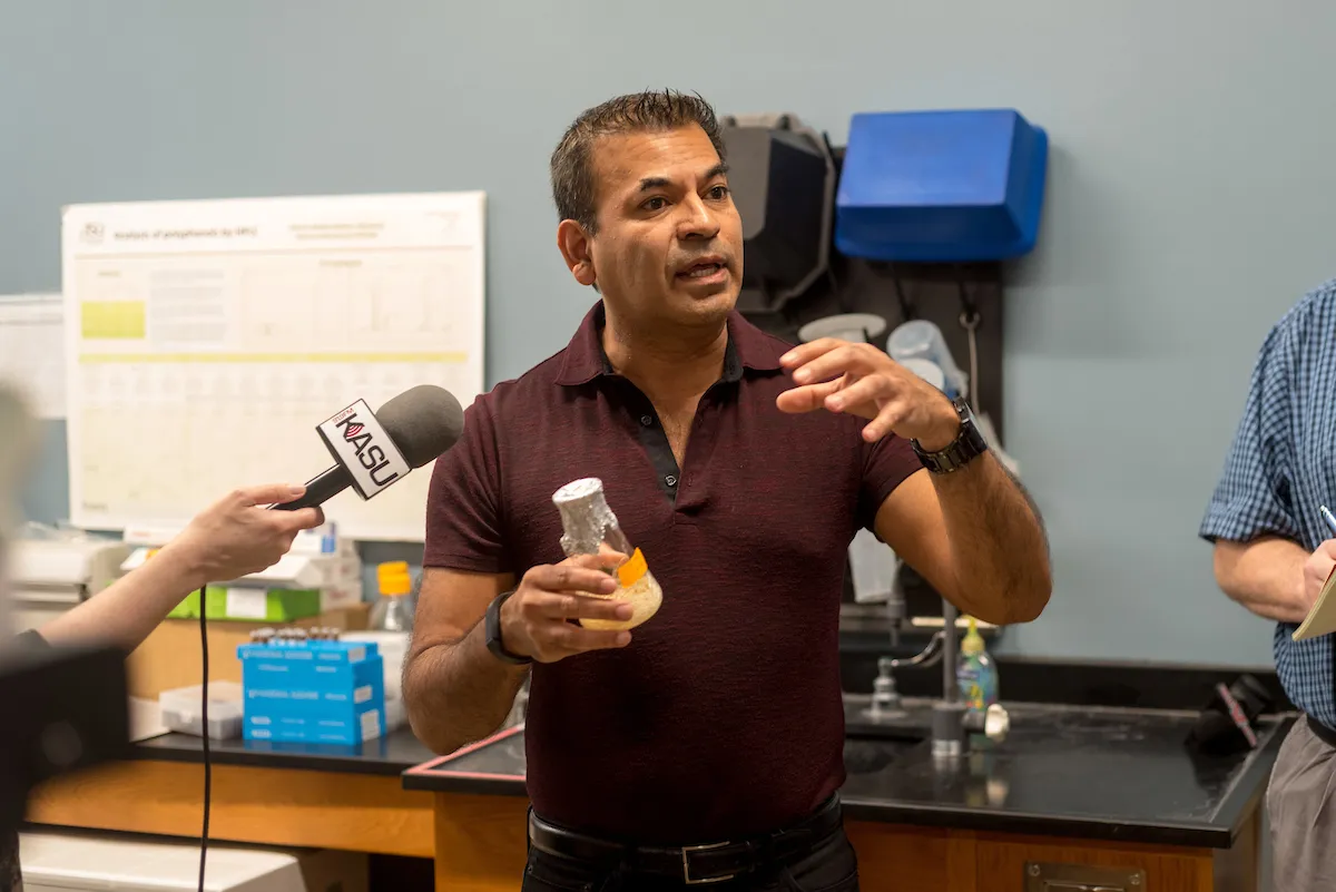 A faculty member holds a lab sample while speaking to a KASU reporter during a research interview.