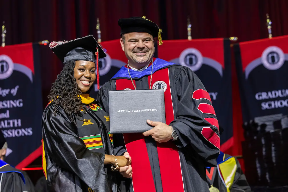 Graduate smiles while receiving diploma during Arkansas State University commencement ceremony with academic banners in the background.