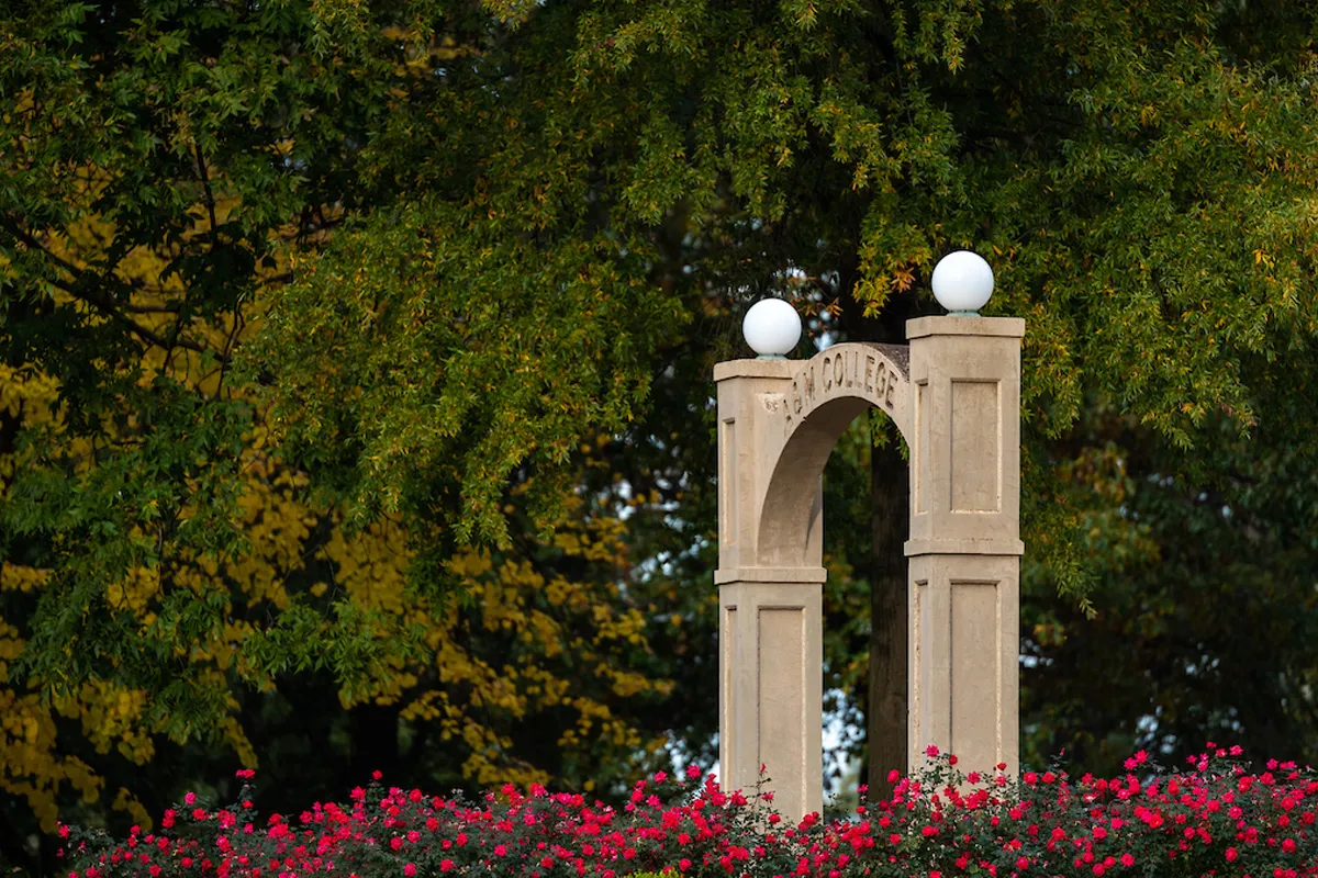 Historic archway from A-State's early years framed by lush trees and blooming flowers on a fall day.