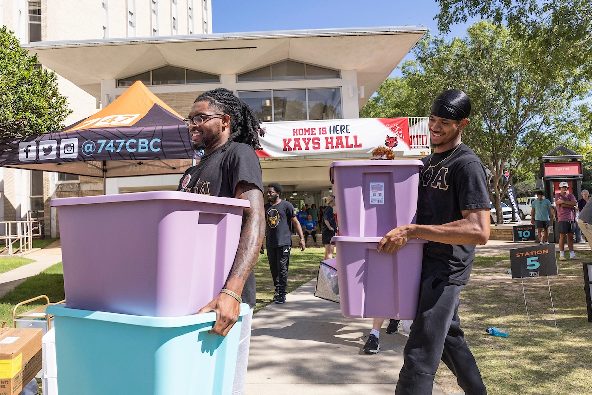 Students wearing Greek letter t-shirts, carrying boxes into residence hall with tents set up outside.