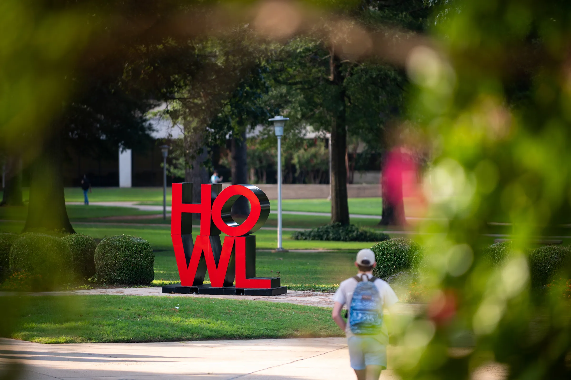 Student walks near bold red HOWL sculpture on a green campus quad with sunny trees in background.