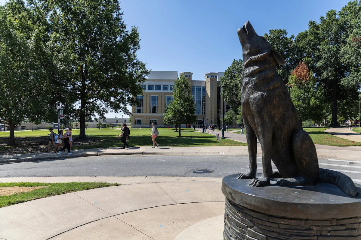 Howl the wolf statue stands in the foreground as students walk past a large campus building on a sunny day.