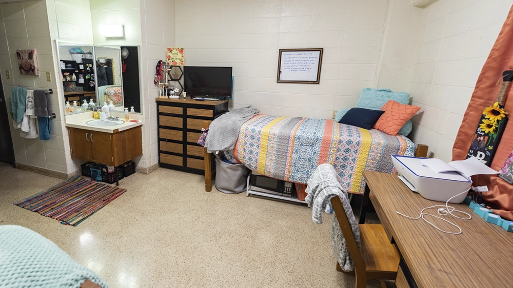 Bedroom with colorful bedding, dresser, and sink in Kays Hall suite.