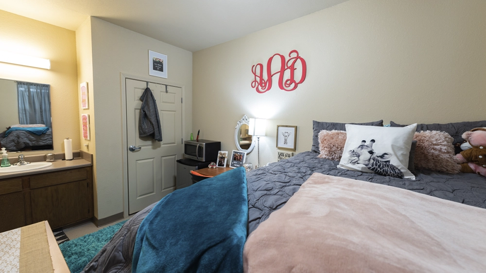 Bedroom with twin bed, floral bedding, desk with chair, mini fridge, and wall collage of photos.
