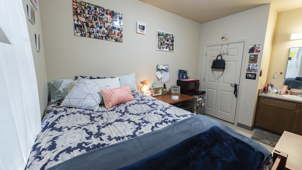 Bedroom with twin bed, floral bedding, desk with chair, and wall photo collage.