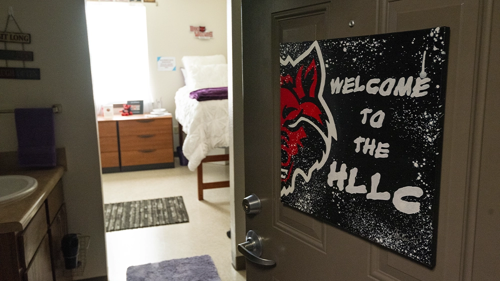 View from bathroom into bedroom with twin bed and wooden dresser, featuring a sign that says ‘Welcome to the HLLC’ on the door.