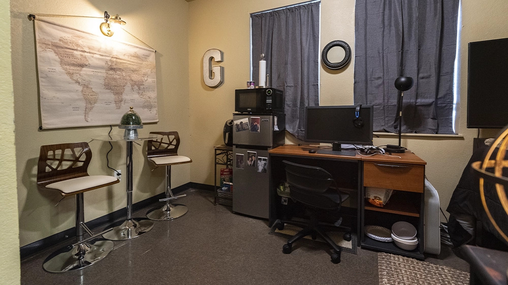 Bedroom with raised bed, black bedding, desk with computer, and two windows with dark curtains.