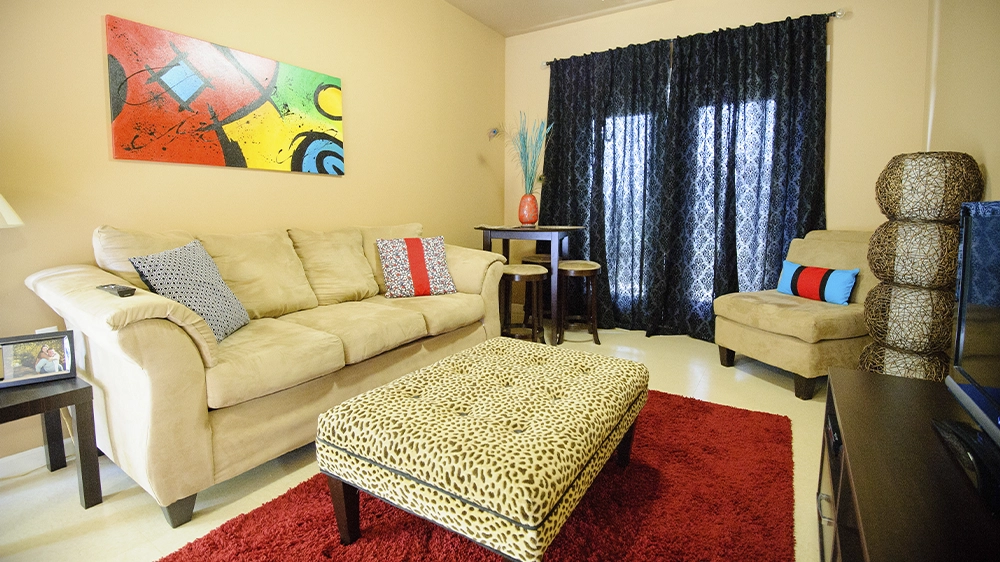 Living room with beige sofa and armchair accented by colorful pillows, leopard-print ottoman on a red rug, small round dining table with two stools, and large patterned curtains behind glass doors. A bright abstract painting hangs on the wall.