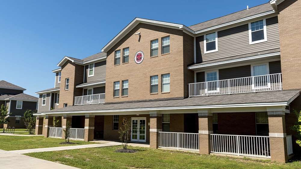 Front view of ROTC Living Learning Community building with tan brick exterior, gray siding, and white railings on covered balconies, set against a clear blue sky.