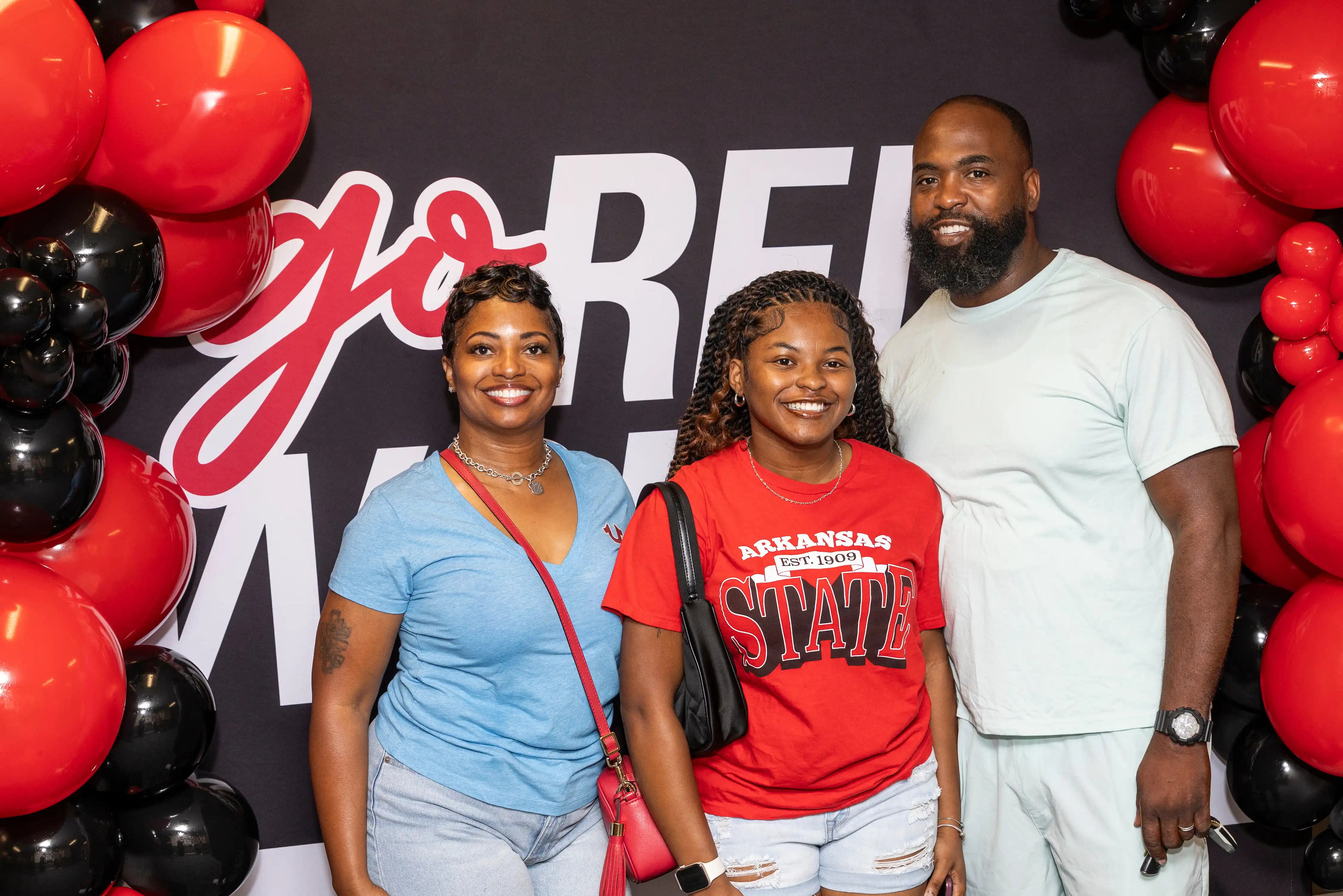 Student with parents in front of red and black backdrop and balloons.