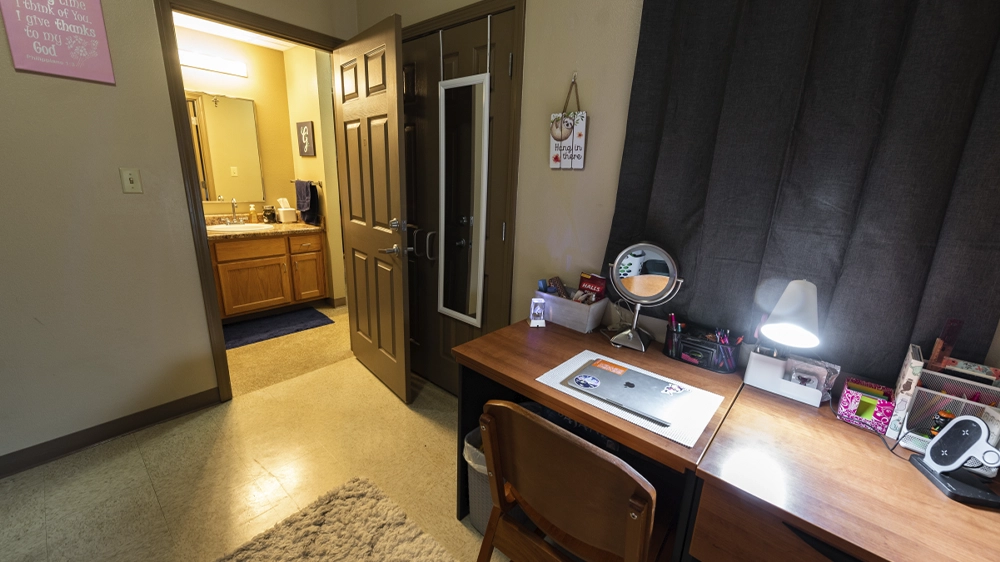Bedroom view showing desk area and open door leading to shared bathroom with sink.