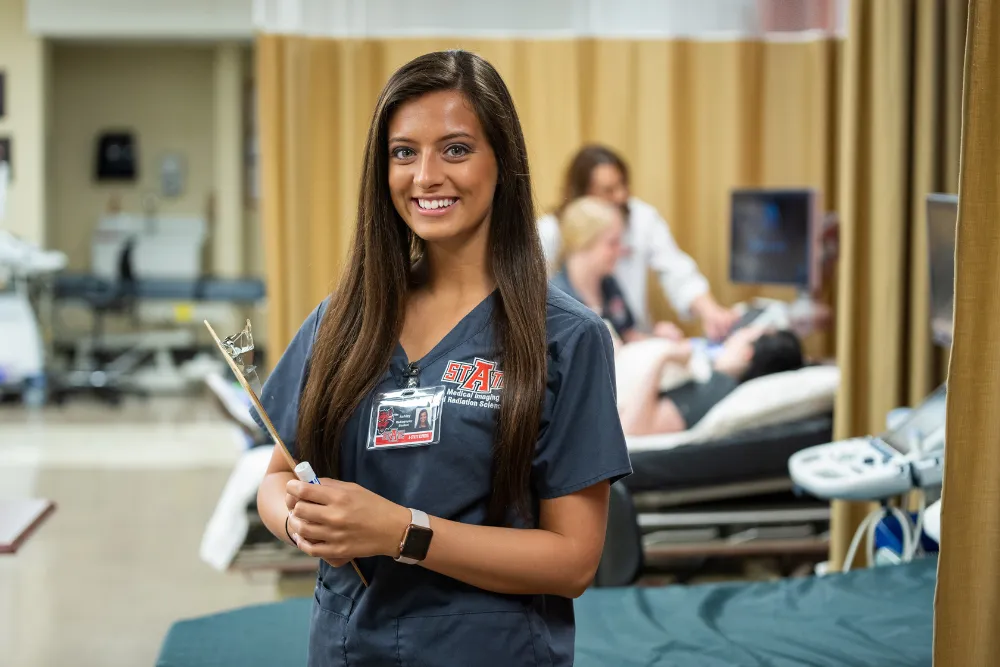 A-State radiology student in clinic holding a clip-board in a-state scrubs.
