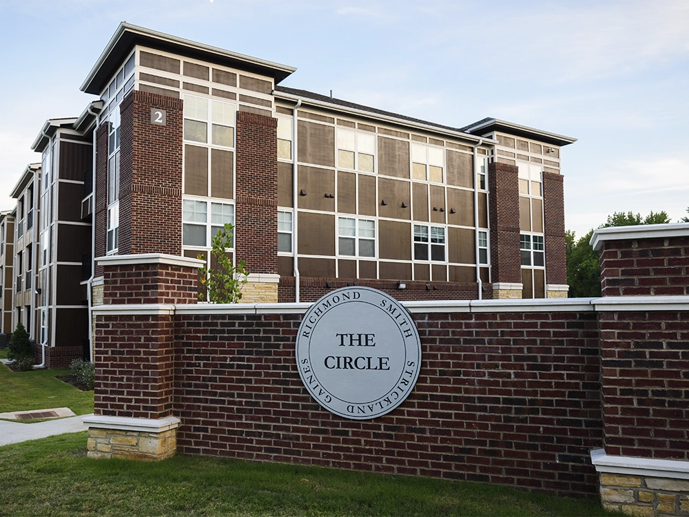 Brick entrance sign for The Circle apartments featuring a circular plaque with the name, in front of a multi-story brick building with large windows.