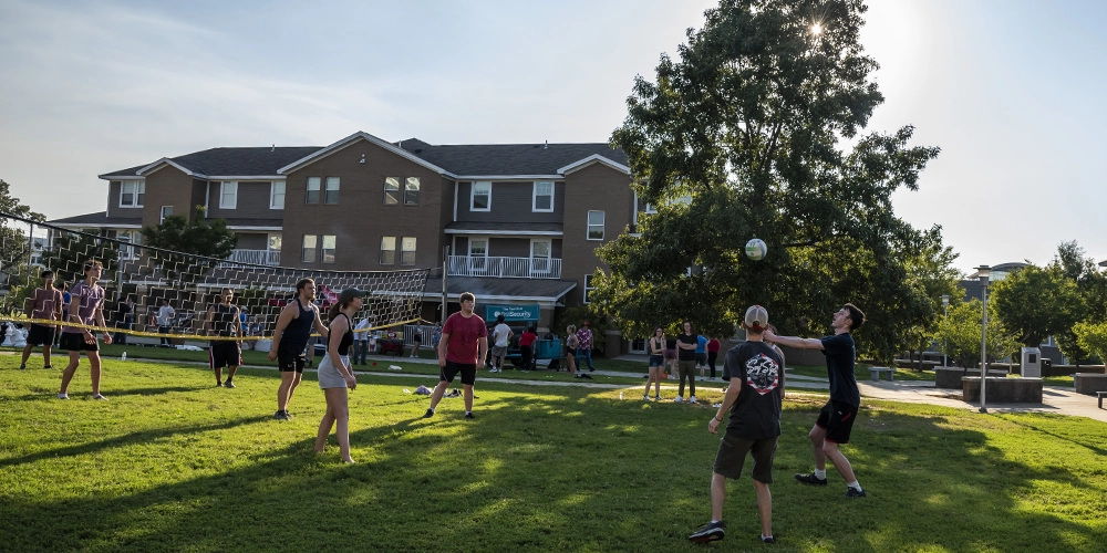 Group of students playing volleyball on a grassy outdoor court with a net, in front of a multi-story apartment-style building on a sunny day.