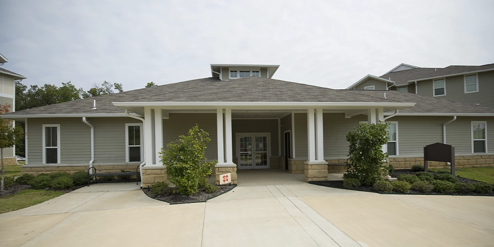 Front entrance of the Red Wolf Den commons building featuring a covered porch with white columns, light gray siding, and landscaped shrubs along the walkway. The building sits on a concrete driveway under a partly cloudy sky.