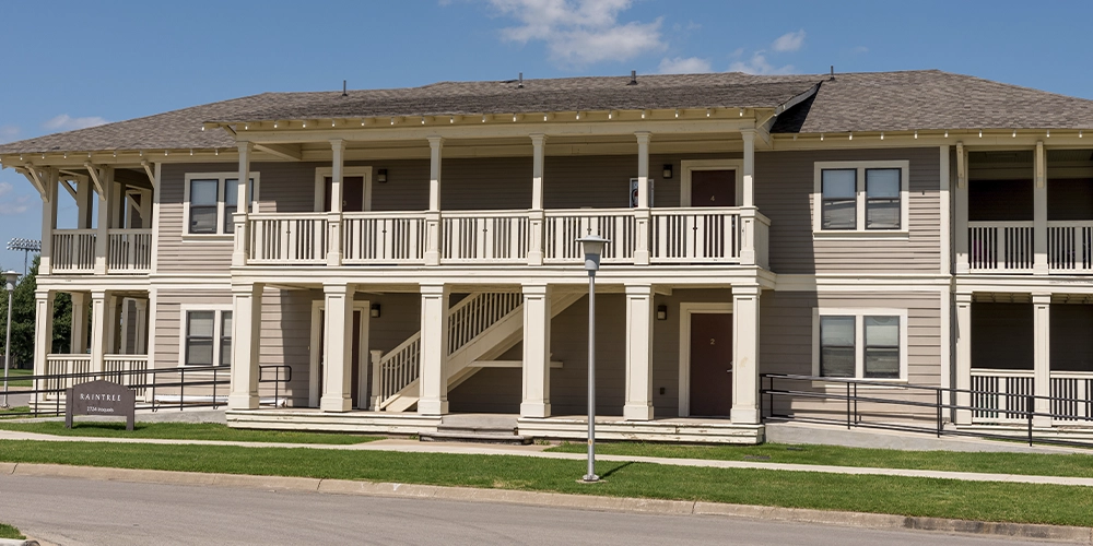 Front view of a two-story beige apartment building at The Village with wide covered balconies, white railings, and a central staircase leading to the upper level, set along a paved street with green grass and a clear blue sky.