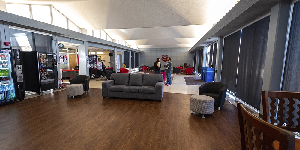 Spacious lobby inside University Hall featuring a gray sofa, round ottomans, vending machines, and large windows with black shades. Several people are visible near the back of the room, and the space includes bright overhead lighting and wood-style flooring.