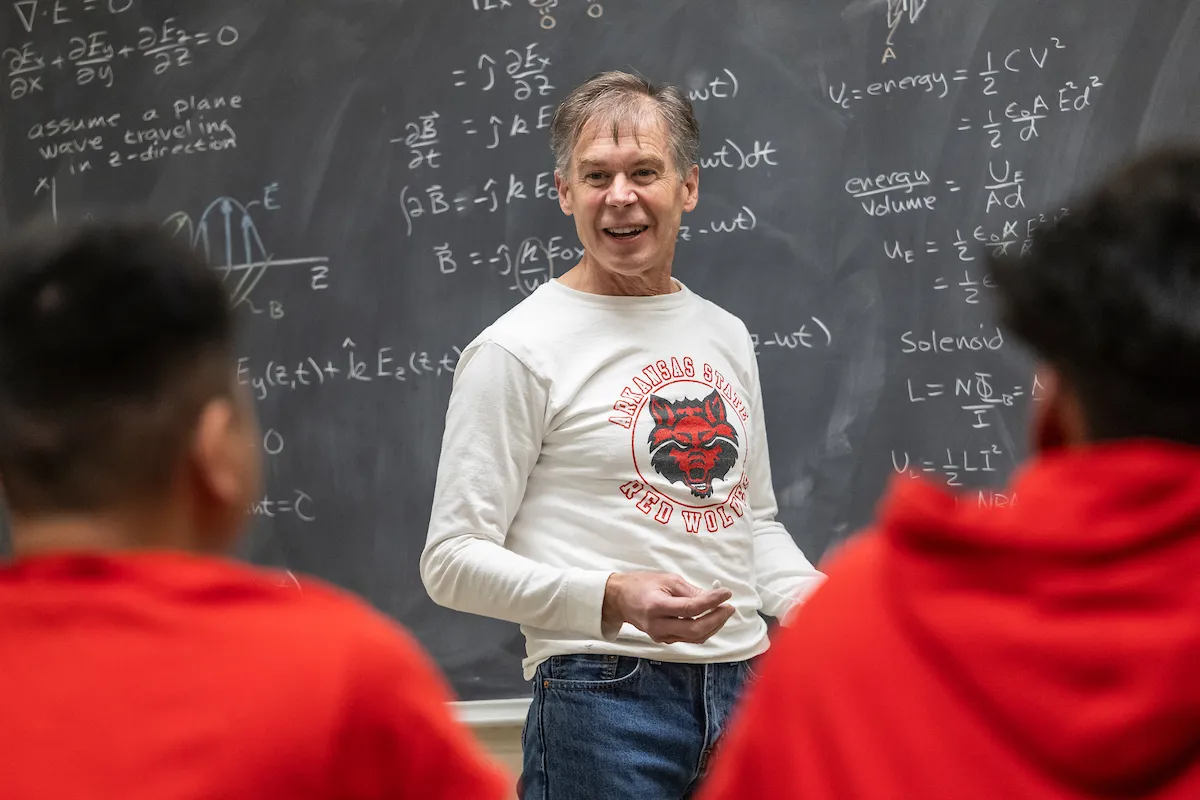 A physics professor stands in front of a chalkboard covered in equations, teaching students.