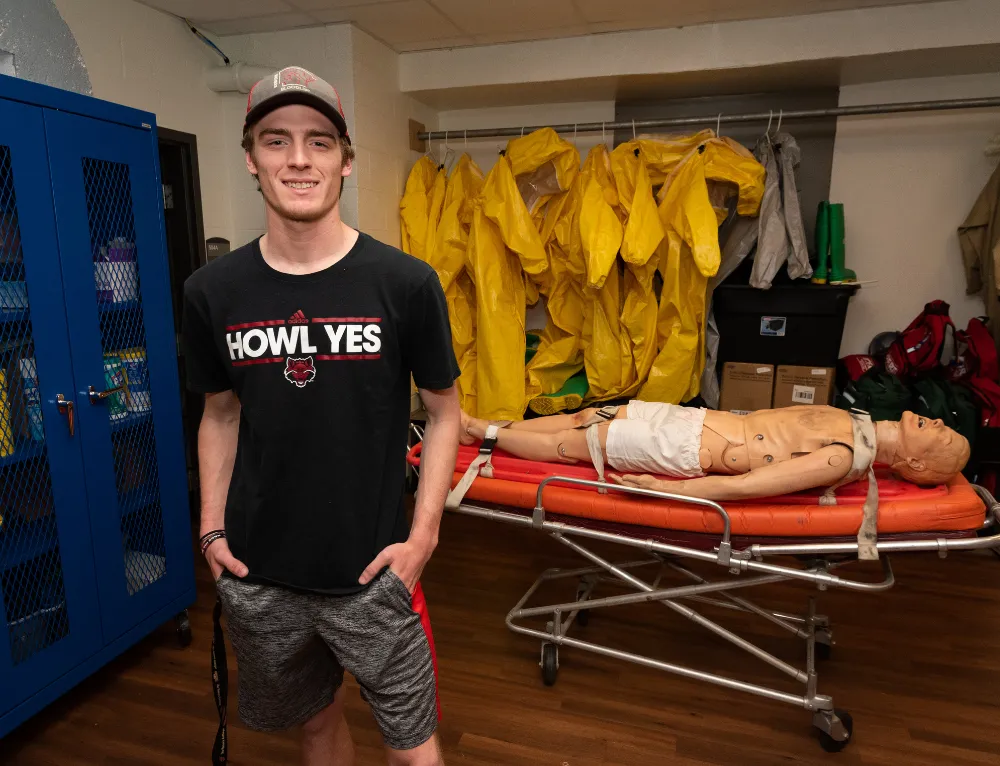 disaster preparedness student posing in a redwolf shirt with a dummy in the background on a stretcher