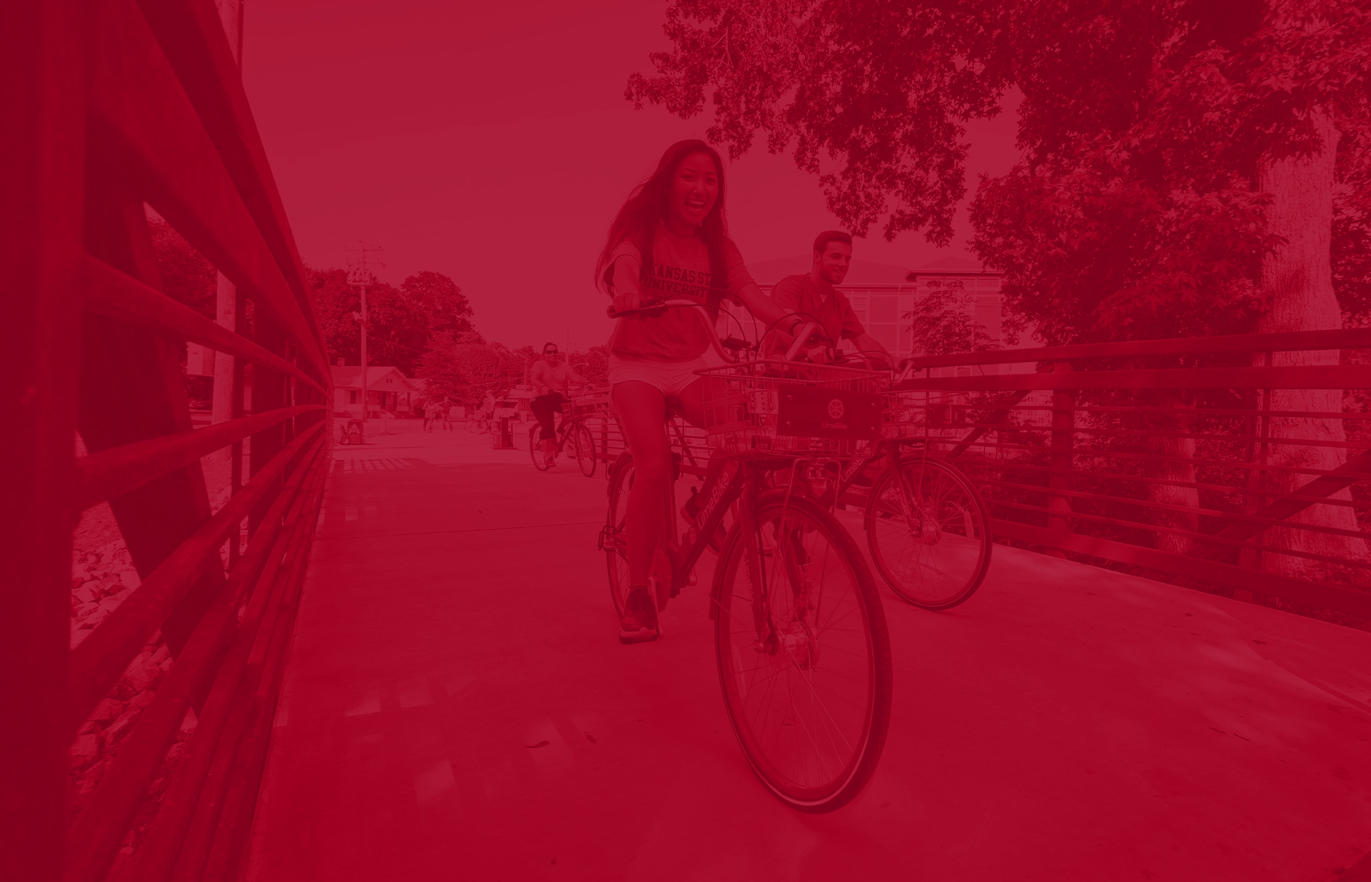 Cyclists riding across a wooden pedestrian bridge on campus, with trees and buildings in the background, shown with a red color overlay.