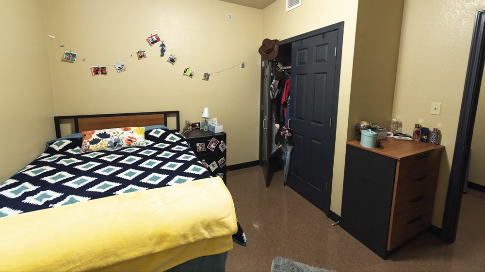 Bedroom view showing full-size bed with patterned blanket, dresser, open closet, and decorative string lights with photos.