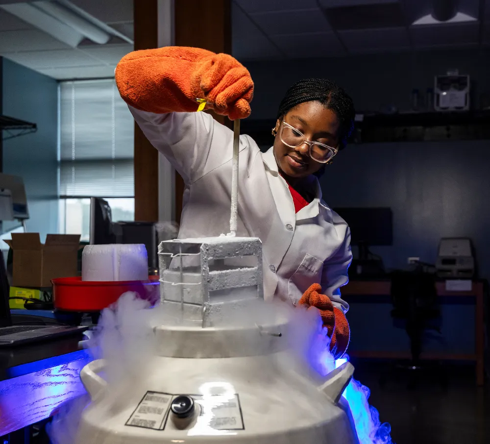 female student wearing a labcoat, safety glasses and heavy orange gloves is using a rope to lift an object from a smoking metal cylinder