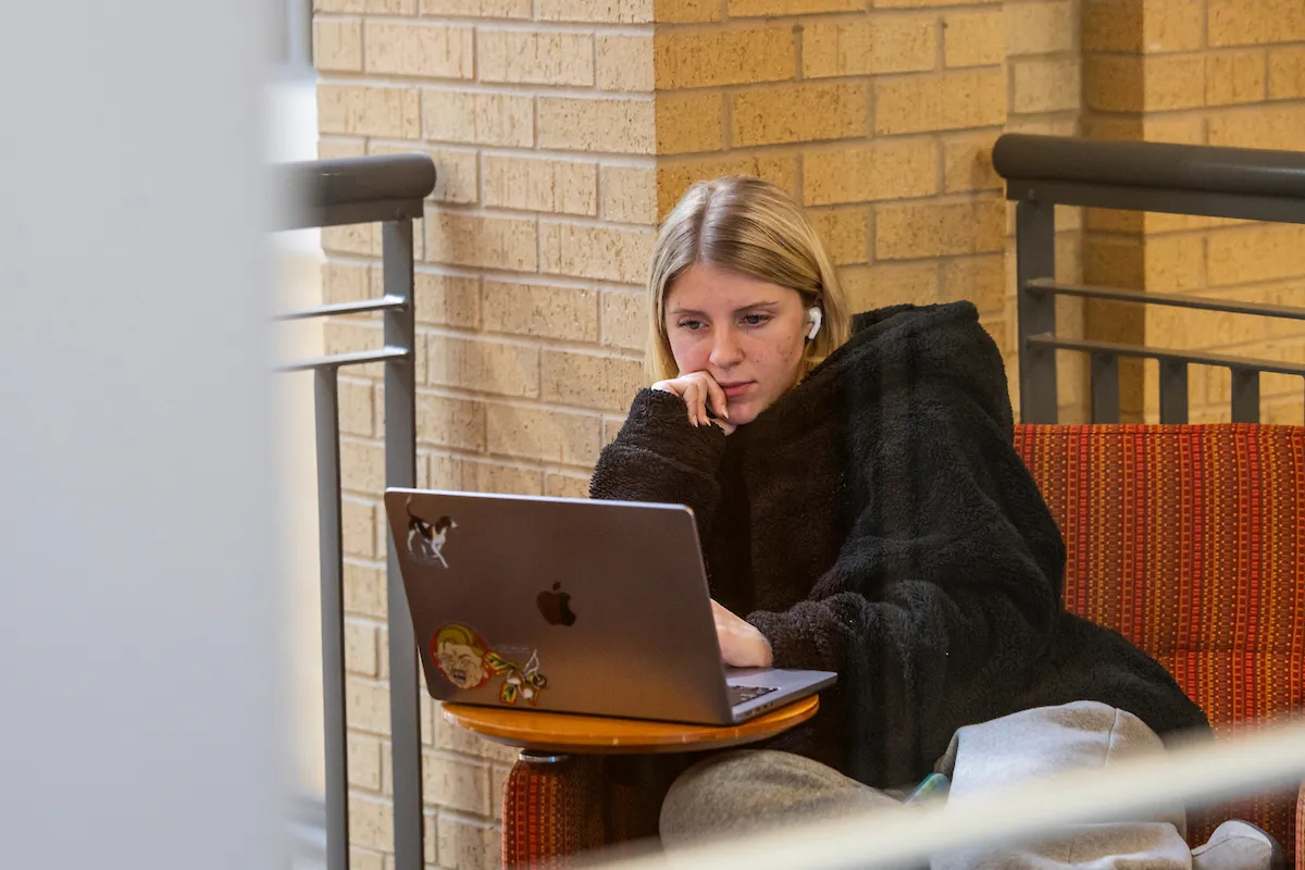 A student lounges in a chair on campus while working on a laptop.