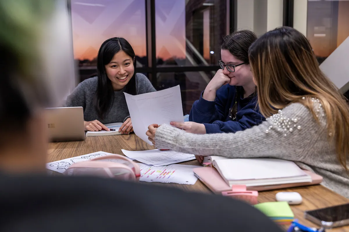 Group of students collaborate on homework at a shared table with notes, laptops, and sunset in the background.